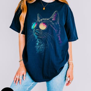 May include: A navy blue t-shirt with a graphic of a cat wearing sunglasses. The cat's eyes are outlined in pink and blue, and the sunglasses have a rainbow gradient.