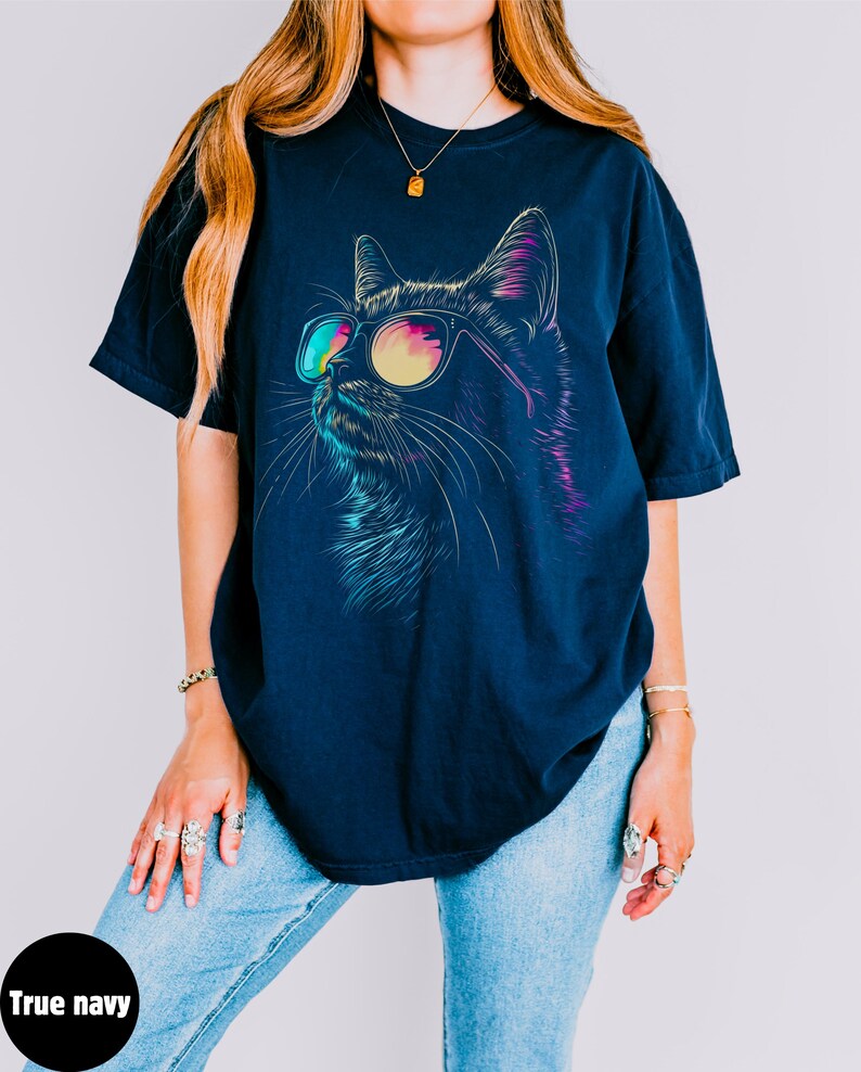 May include: A navy blue t-shirt with a graphic of a cat wearing sunglasses. The cat's eyes are outlined in pink and blue, and the sunglasses have a rainbow gradient.