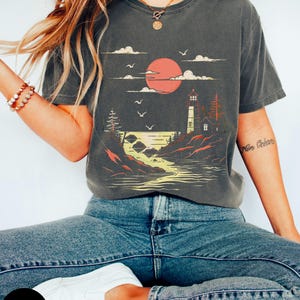May include: A dark gray t-shirt with a graphic print of a lighthouse on a rocky shore with a red sun setting behind it. The text "More Oceans" is printed on the right sleeve.
