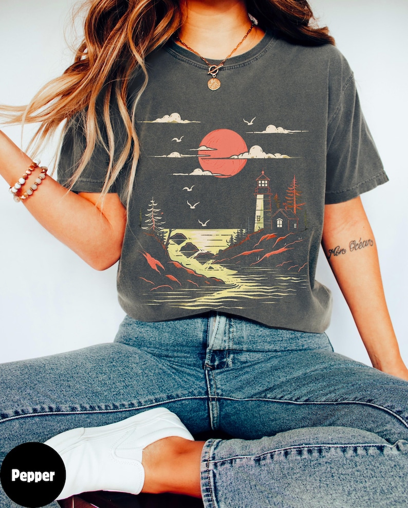 May include: A dark gray t-shirt with a graphic print of a lighthouse on a rocky shore with a red sun setting behind it. The text "More Oceans" is printed on the right sleeve.