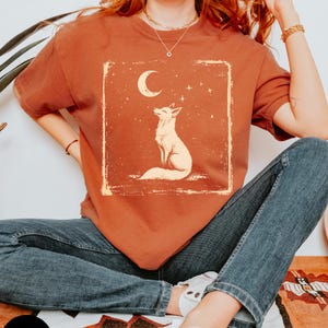 May include: A rust-colored t-shirt featuring a cream-colored illustration of a fox gazing at a crescent moon and stars. The design is within a rectangular frame with a distressed, vintage aesthetic. The shirt is paired with blue jeans.