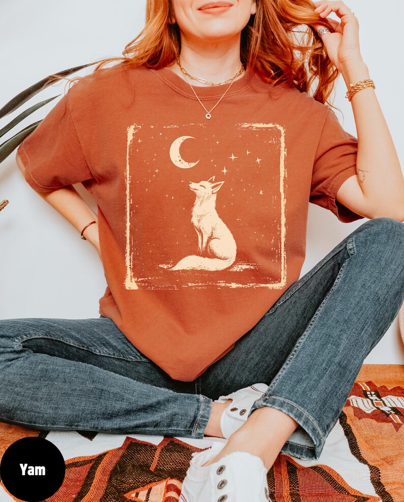 May include: A rust-colored t-shirt featuring a cream-colored illustration of a fox gazing at a crescent moon and stars. The design is within a rectangular frame with a distressed, vintage aesthetic. The shirt is paired with blue jeans.