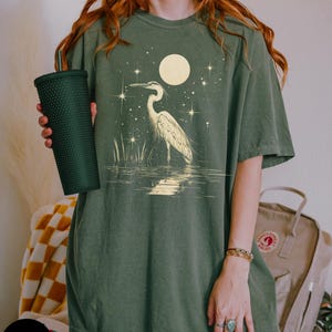 Puede incluir: Una camiseta verde de gran tama&ntilde;o con un gr&aacute;fico de garza blanca de pie en el agua con una luna llena y estrellas en el fondo. El texto "Moss" es visible en la esquina inferior izquierda de la imagen.