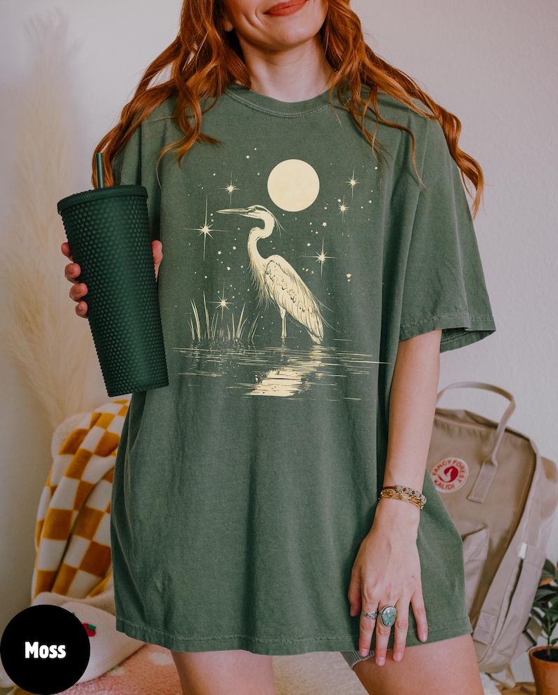 Puede incluir: Una camiseta verde de gran tama&ntilde;o con un gr&aacute;fico de garza blanca de pie en el agua con una luna llena y estrellas en el fondo. El texto "Moss" es visible en la esquina inferior izquierda de la imagen.