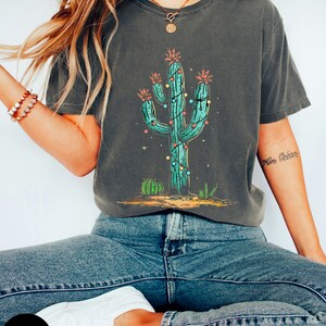 以下が含まれることがあります： 緑色のサボテンにカラフルなクリスマスライトが飾られたダークグレーのTシャツ。サボテンの周りは小さなサボテンと星で囲まれています。Tシャツはゆったりとしたフィット感で、袖は短いです。