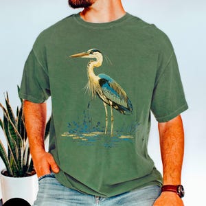 May include: A moss green t-shirt featuring a blue and white heron graphic. The heron is standing in a pond with reeds.