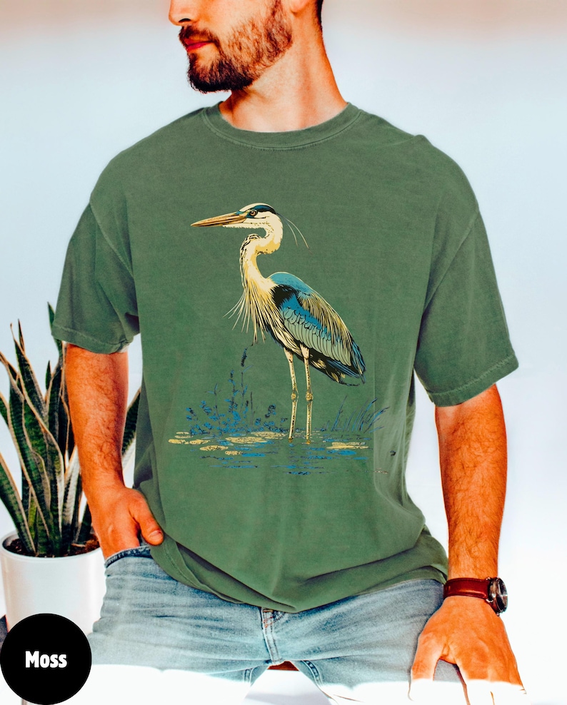 May include: A moss green t-shirt featuring a blue and white heron graphic. The heron is standing in a pond with reeds.