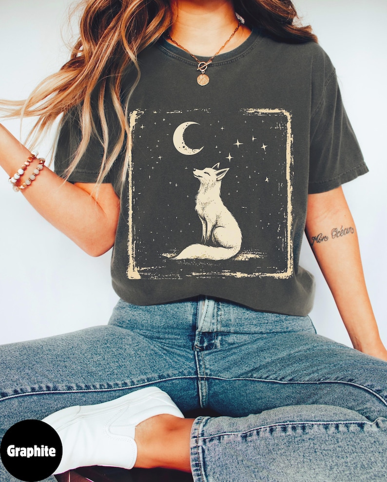 May include: Graphite-coloured t-shirt with a cream illustration of a fox looking at a crescent moon and stars, framed in a rectangle. The shirt is worn with blue jeans and white trainers.