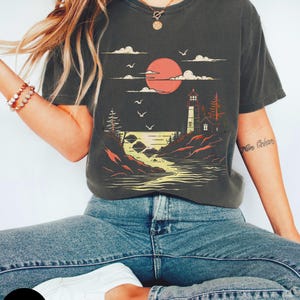 May include: A graphite grey t-shirt with a graphic print of a lighthouse on a rocky shore with a red sun setting behind it. The graphic also includes clouds and birds flying in the sky. The text "Min Oc&eacute;ans" is visible on the right sleeve of the shirt.