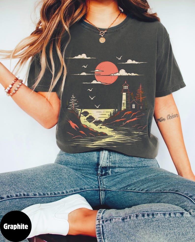 May include: A graphite grey t-shirt with a graphic print of a lighthouse on a rocky shore with a red sun setting behind it. The graphic also includes clouds and birds flying in the sky. The text "Min Oc&eacute;ans" is visible on the right sleeve of the shirt.