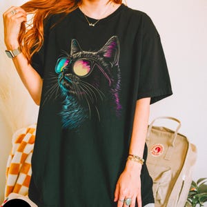 May include: Black oversized t-shirt featuring a graphic of a cat wearing sunglasses. The cat's fur is outlined in blue and purple.