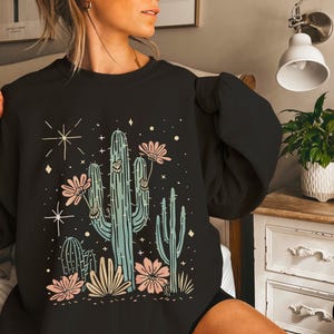 May include: Black sweatshirt with a graphic print of a cactus and flowers. The cactus is green and the flowers are pink and white.