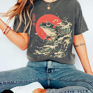 Pode incluir: Uma camiseta cinza com uma estampa gr&aacute;fica de um sapo verde em uma onda com um sol vermelho atr&aacute;s. O texto japon&ecirc;s na camiseta diz "波乗り蛙".