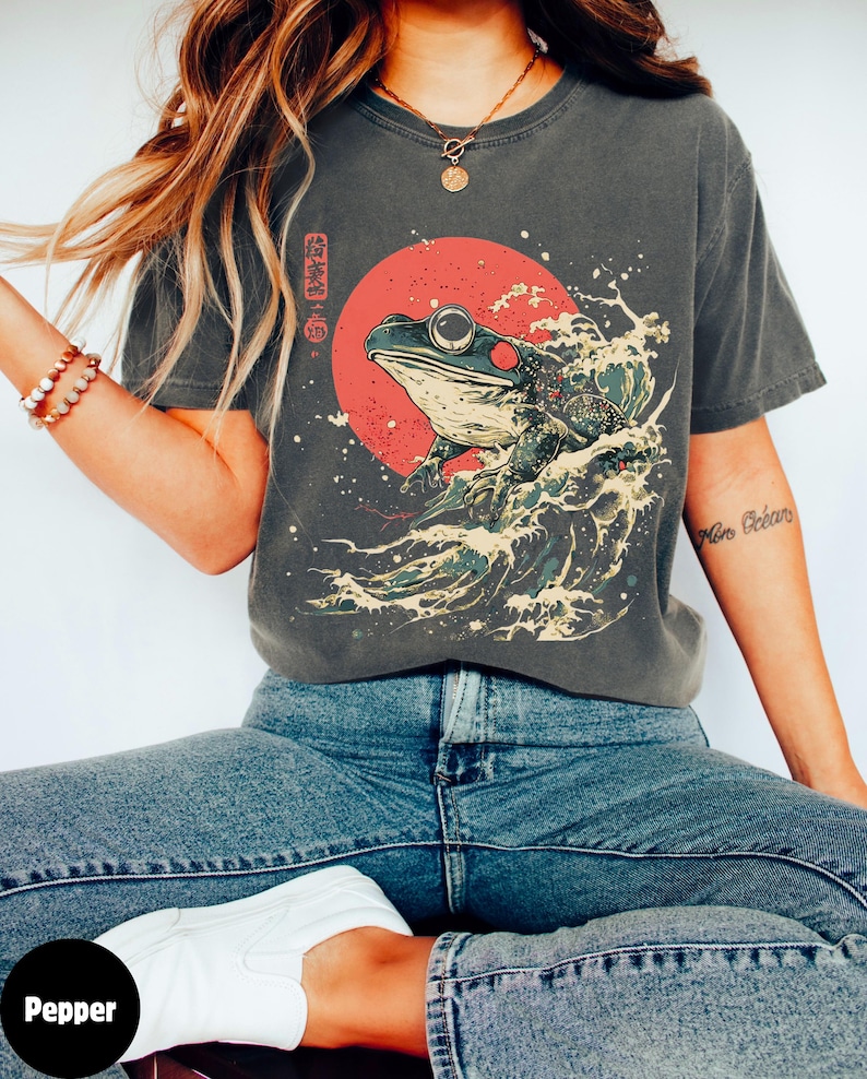 Pode incluir: Uma camiseta cinza com uma estampa gr&aacute;fica de um sapo verde em uma onda com um sol vermelho atr&aacute;s. O texto japon&ecirc;s na camiseta diz "波乗り蛙".