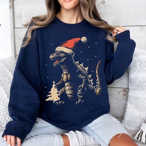 Può includere: Una felpa blu navy con una grafica di un dinosauro che indossa un cappello da Babbo Natale e sta in piedi davanti a un albero di Natale. Il dinosauro è verde e ha un cappello da Babbo Natale rosso con bordatura bianca. Lo sfondo è blu scuro con fiocchi di neve bianchi.