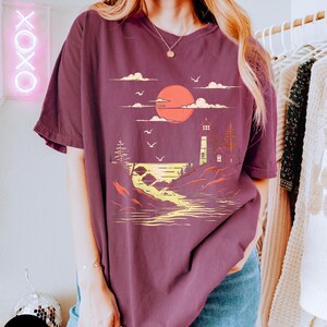 May include: A maroon oversized t-shirt with a graphic print of a lighthouse, a sunset, and a body of water. The graphic is in yellow, orange, and white.