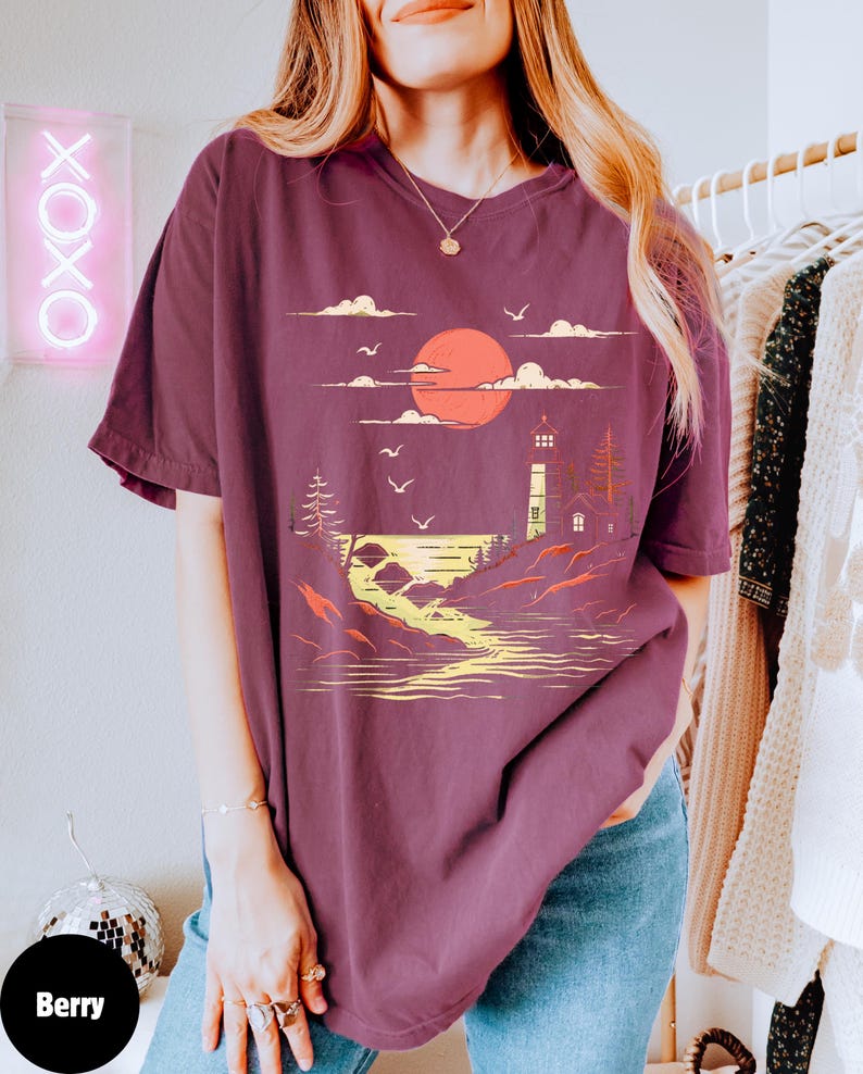 May include: A maroon oversized t-shirt with a graphic print of a lighthouse, a sunset, and a body of water. The graphic is in yellow, orange, and white.
