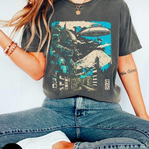 Könnte beinhalten: Ein dunkelgraues T-Shirt mit einem grafischen Druck eines Roboters, einer fliegenden Untertasse und einer Stadtlandschaft. Die Grafik ist in Blautönen, Weiß und Schwarz gehalten. Das T-Shirt hat einen Rundhalsausschnitt und kurze Ärmel.