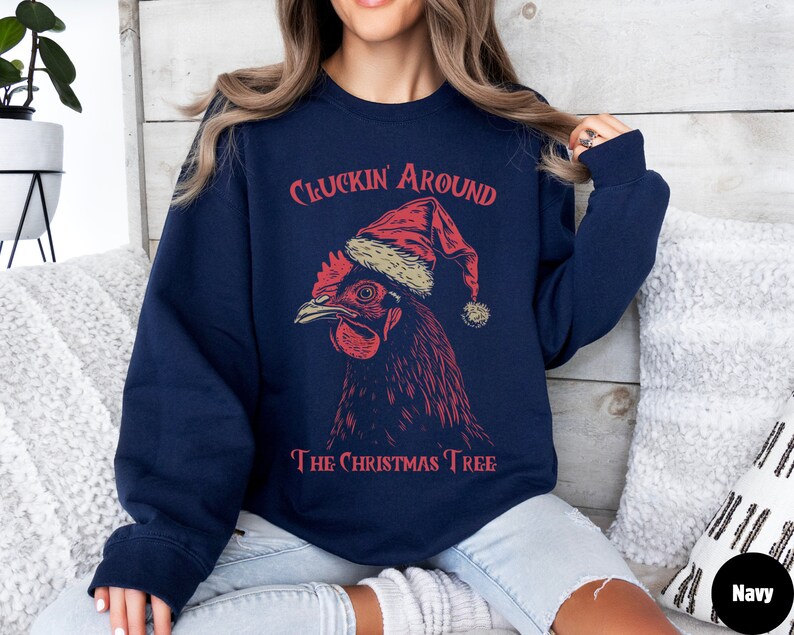 May include: A navy blue sweatshirt featuring a red and white illustration of a chicken wearing a Santa hat. The text "Cluckin' Around The Christmas Tree" is printed on the sweatshirt.