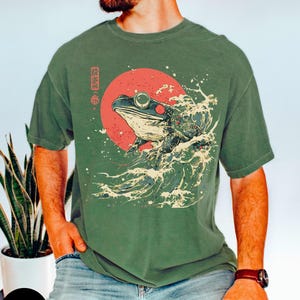 Pode incluir: Uma camiseta verde com um gr&aacute;fico de um sapo em uma onda com um sol vermelho atr&aacute;s. O texto japon&ecirc;s na camiseta diz "蛙波".