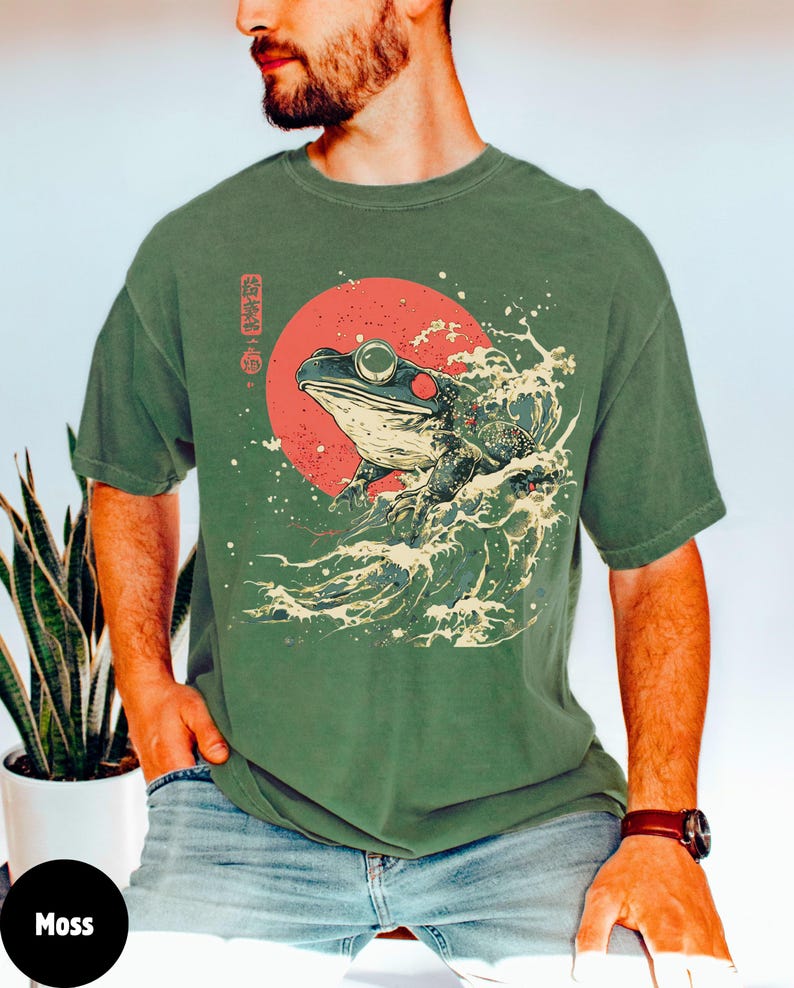 Pode incluir: Uma camiseta verde com um gr&aacute;fico de um sapo em uma onda com um sol vermelho atr&aacute;s. O texto japon&ecirc;s na camiseta diz "蛙波".