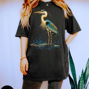 May include: A black t-shirt featuring a graphic of a blue heron standing in water. The heron has a long neck and long legs. The graphic is in a vintage style.