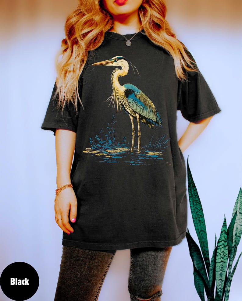 May include: A black t-shirt featuring a graphic of a blue heron standing in water. The heron has a long neck and long legs. The graphic is in a vintage style.