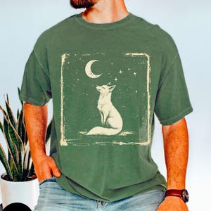 May include: Moss green t-shirt featuring a white fox illustration under a crescent moon and stars. The design is framed within a distressed rectangle. The shirt is a relaxed fit.