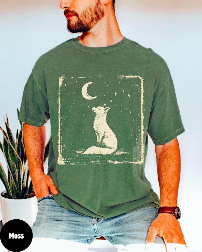 May include: Moss green t-shirt featuring a white fox illustration under a crescent moon and stars. The design is framed within a distressed rectangle. The shirt is a relaxed fit.
