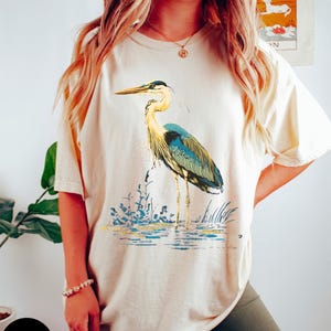 May include: A cream coloured t-shirt with a blue and green heron graphic. The heron is standing in water with green reeds. The shirt is labelled "Ivory".