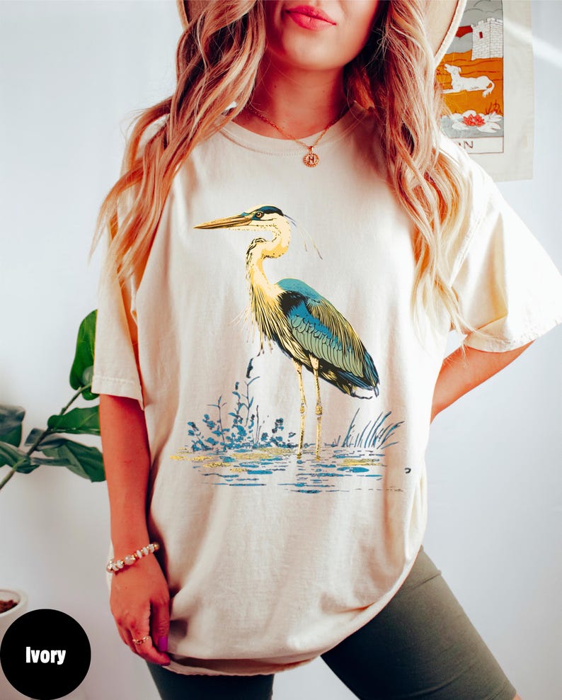 May include: A cream coloured t-shirt with a blue and green heron graphic. The heron is standing in water with green reeds. The shirt is labelled "Ivory".