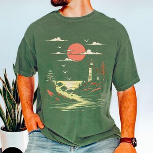 May include: A green t-shirt with a graphic of a lighthouse, a red sun, and a river flowing into the ocean. The graphic is in a vintage style.