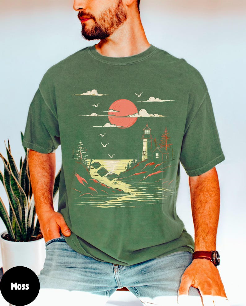 May include: A green t-shirt with a graphic of a lighthouse, a red sun, and a river flowing into the ocean. The graphic is in a vintage style.