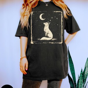 May include: Black t-shirt featuring a white fox illustration under a crescent moon and stars. The design is framed in a rectangular border. The shirt has short sleeves and a relaxed fit, suitable for casual wear.