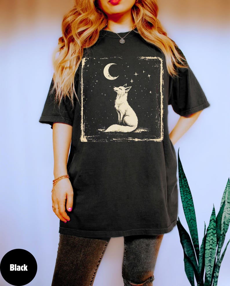 May include: Black t-shirt featuring a white fox illustration under a crescent moon and stars. The design is framed in a rectangular border. The shirt has short sleeves and a relaxed fit, suitable for casual wear.