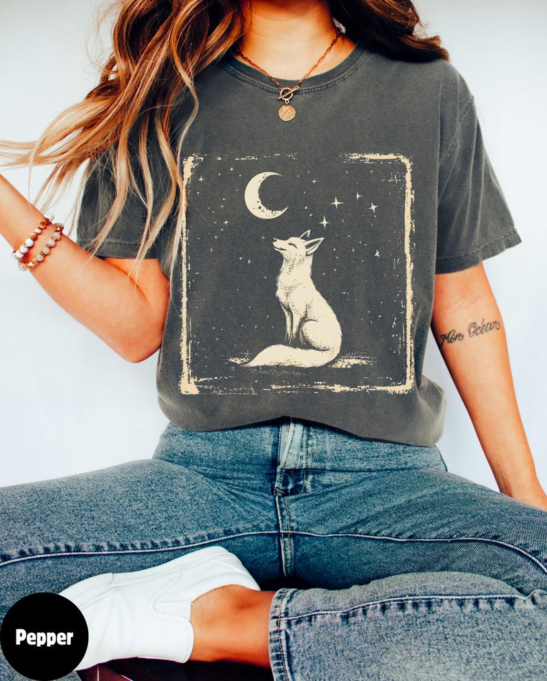 May include: Dark gray t-shirt featuring a white fox gazing at a crescent moon and stars within a rectangular frame. The shirt is a crew neck and has short sleeves. The word "Pepper" is in a black circle in the bottom left corner.