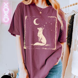 May include: Berry-colored t-shirt featuring a cream-colored fox design under a crescent moon and stars. The graphic is within a rectangular frame. The shirt has short sleeves and a relaxed fit. The word "Berry" is on a round sticker.