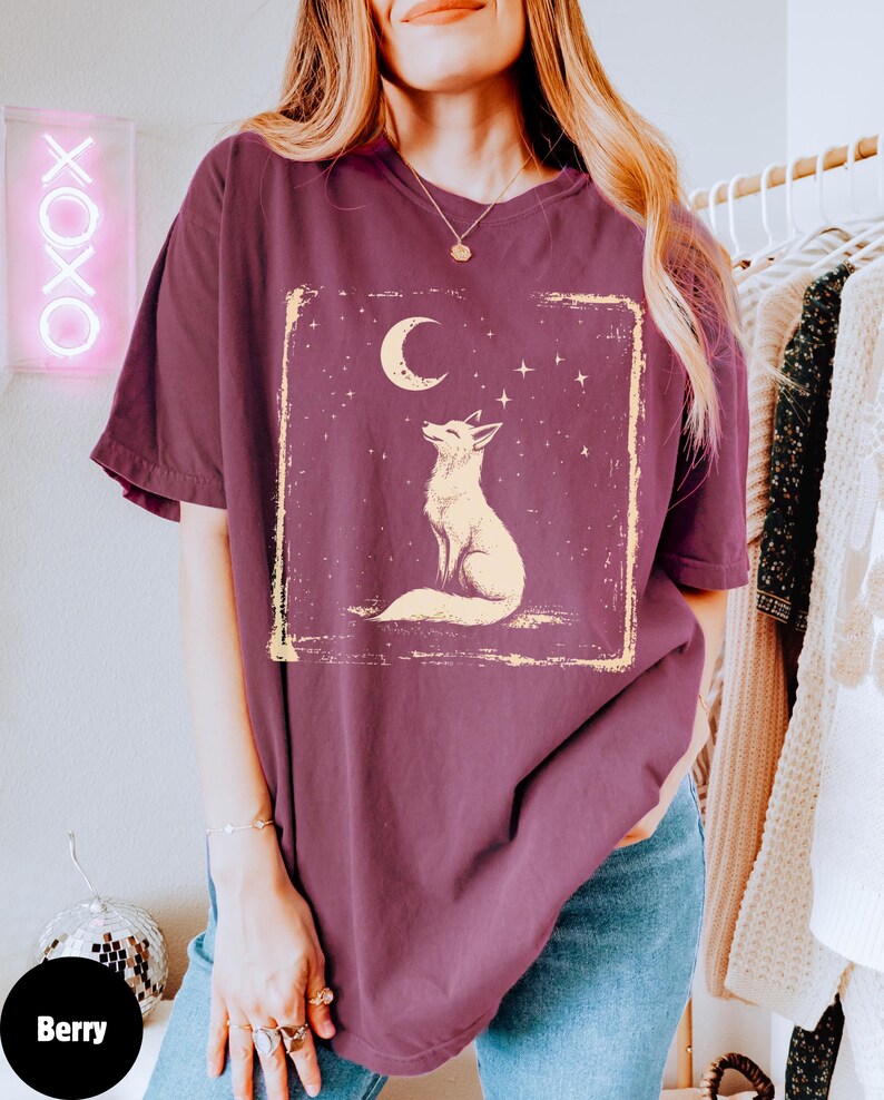 May include: Berry-colored t-shirt featuring a cream-colored fox design under a crescent moon and stars. The graphic is within a rectangular frame. The shirt has short sleeves and a relaxed fit. The word "Berry" is on a round sticker.
