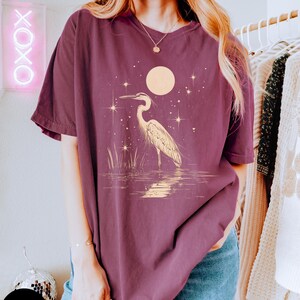 Puede incluir: Una camiseta de gran tama&ntilde;o de color marr&oacute;n con un gr&aacute;fico de una garza blanca de pie en un cuerpo de agua con una luna llena y estrellas en el fondo.