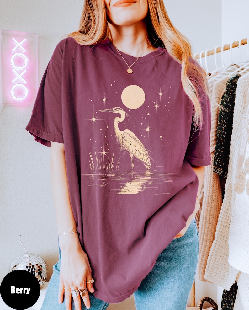 Puede incluir: Una camiseta de gran tama&ntilde;o de color marr&oacute;n con un gr&aacute;fico de una garza blanca de pie en un cuerpo de agua con una luna llena y estrellas en el fondo.