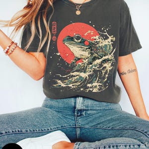 Pode incluir: Uma camiseta cinza grafite com uma estampa gr&aacute;fica de um sapo sentado em uma onda com um sol vermelho atr&aacute;s. O texto japon&ecirc;s na camiseta diz "蛙"