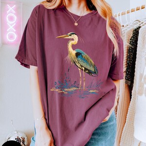 May include: An oversized maroon t-shirt featuring a graphic of a blue heron standing in a pond. The heron has a long neck and long legs. The heron is standing in a pond with green reeds.