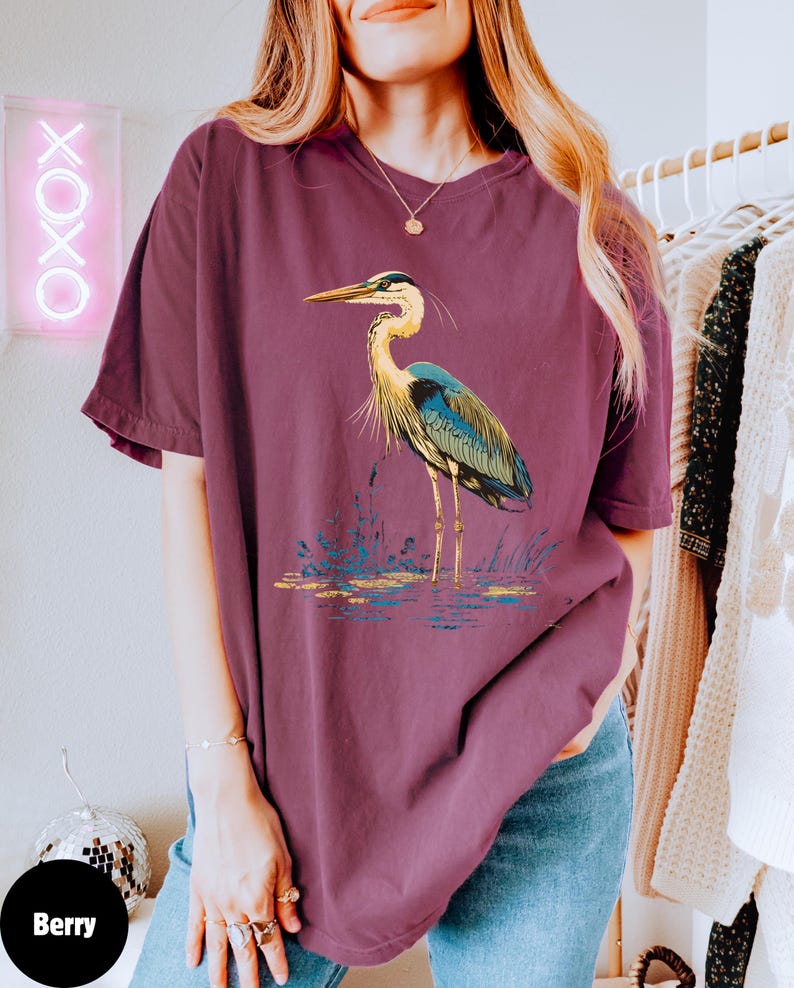 May include: An oversized maroon t-shirt featuring a graphic of a blue heron standing in a pond. The heron has a long neck and long legs. The heron is standing in a pond with green reeds.