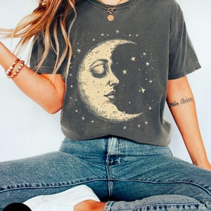 Op de afbeelding: Een antracietgrijs T-shirt met een vintage illustratie van een halve maan met een gezicht en sterren. De tekst "More Dreams" is zichtbaar op de rechtermouw.