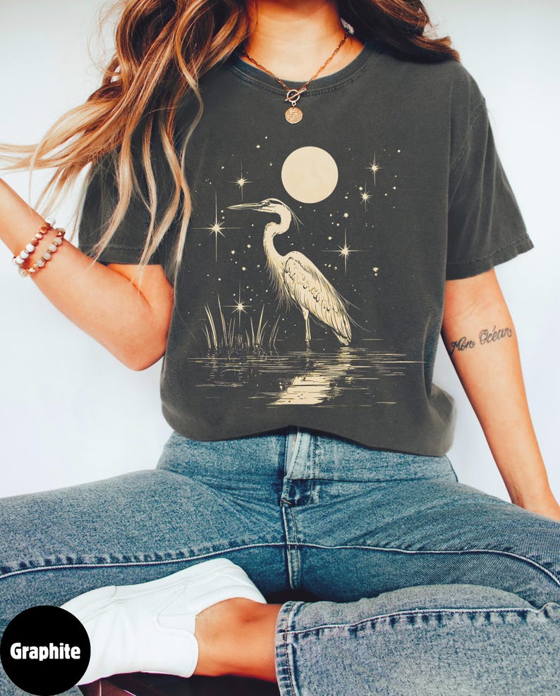 Puede incluir: Una camiseta gris grafito con una garza blanca de pie en un cuerpo de agua bajo una luna llena y estrellas. El texto "Min Oc&eacute;ans" es visible en la manga derecha.