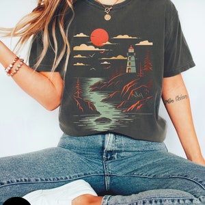 May include: A graphite grey t-shirt with a graphic print of a lighthouse on a rocky shore with a red sun setting in the background. The graphic is in a vintage style.