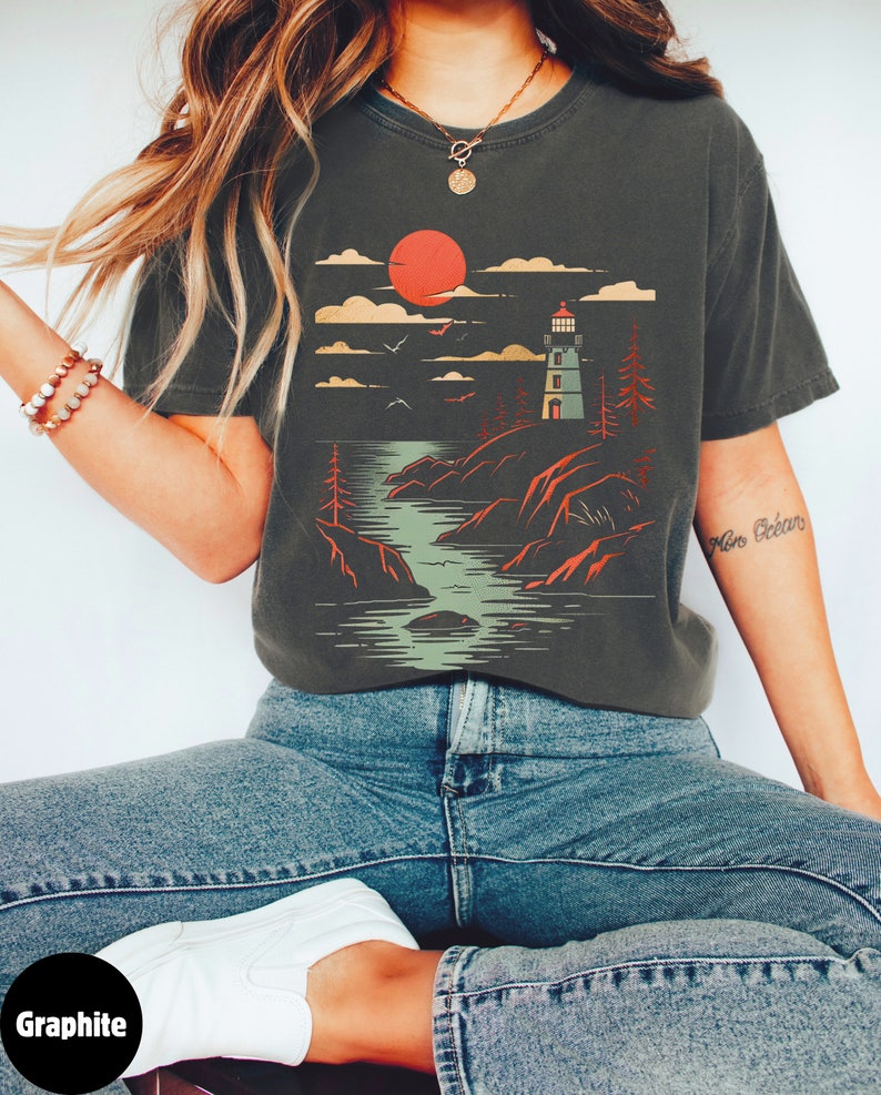 May include: A graphite grey t-shirt with a graphic print of a lighthouse on a rocky shore with a red sun setting in the background. The graphic is in a vintage style.