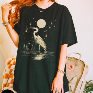 Puede incluir: Una camiseta negra de gran tama&ntilde;o con un estampado gr&aacute;fico de una garza blanca de pie en el agua bajo una luna llena y estrellas.