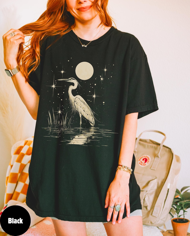 Puede incluir: Una camiseta negra de gran tama&ntilde;o con un estampado gr&aacute;fico de una garza blanca de pie en el agua bajo una luna llena y estrellas.