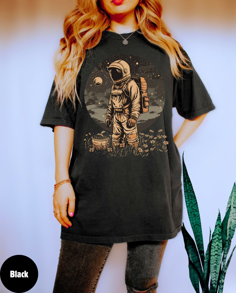 May include: A black t-shirt featuring a graphic of an astronaut standing in a field of flowers. The astronaut is wearing a white spacesuit and holding a bucket. The background is a starry night sky with a planet in the distance.